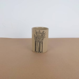 May include: A beige ceramic jar with a black and white illustration of celery stalks in a container. The jar is labeled "CELERY."