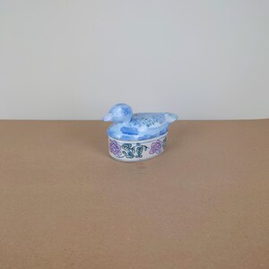 以下が含まれることがあります： 青と白のセラミック製の、アヒルの形をした小箱。蓋が付いており、ピンクと緑の花で飾られています。