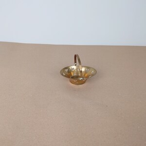May include: A small, gold-coloured metal basket with a handle and heart-shaped cut-outs. The basket is sitting on a brown surface.