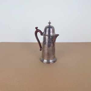May include: Silver coffee pot with a wooden handle and a lid. The pot has a rounded body and a spout.