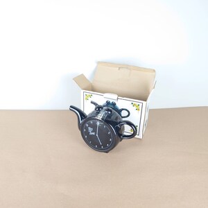 May include: Black ceramic teapot shaped like a clock with the words "Tea Time" on the face. The teapot is in its original box.