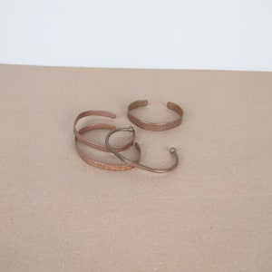 May include: Four copper bracelets with a hammered finish. The bracelets are stacked on top of each other and have a brass accent.