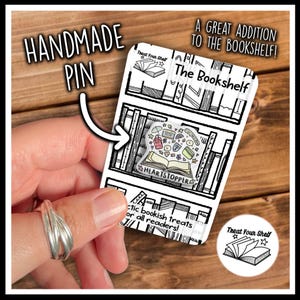 May include: A white card with black outlines of bookshelves and the text "Treat Your Shelf" and "The Bookshelf". A colourful enamel pin with the text "Heartstopper" is shown on the card.