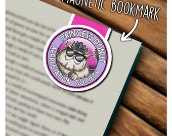 May include: A magnetic bookmark featuring a circular design with a fluffy cat wearing sunglasses and a crown. The text reads PRINCESS DONUT APPRECIATION SOCIETY. The bookmark is pink and purple with a white border, clipped onto a book page.