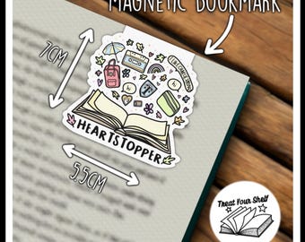 Op de afbeelding: Een witte magnetische bladwijzer met een open boek met de tekst "HEARTSTOPPER" en verschillende illustraties, waaronder een regenboog, een cassettebandje, een rugzak en de tekst "LIKE CHARLIE SPRING". De bladwijzer meet 7 cm bij 5,5 cm.