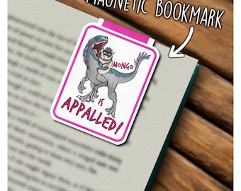 May include: A rectangular magnetic bookmark with a pink border featuring a blue dinosaur holding a small owl wearing glasses. The text reads "MONGO IS APPALLED!" in pink letters. The bookmark is clipped onto the edge of an open book page.