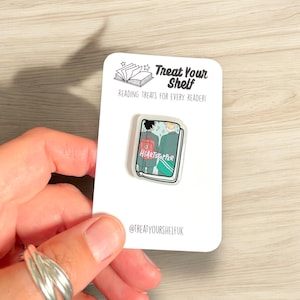 May include: A clear plastic pin featuring a cartoon illustration of a book cover. The book cover depicts two characters, one with black hair and the other with blonde hair. The book title is "Heartstopper". The pin is displayed on a white card with the text "Treat Your Shelf" and "Reading Treats for Every Reader!" The card also includes the text "@treatyourshelfuk".