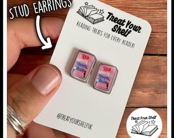 Può includere: Un paio di orecchini a bottone con una copertina di libro rosa e bianca con il titolo "Red, White & Royal Blue". Gli orecchini sono su una carta bianca con il testo "Treat Your Shelf" e "Reading Treats for Every Reader!"