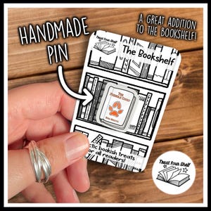 May include: A white enamel pin shaped like a book with an orange paw print graphic and the text The Furry Count, attached to a rectangular backing card featuring a black and white illustration of a bookshelf and the text Treat Your Shelf.