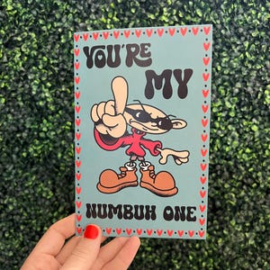 May include: A greeting card with the text "YOU'RE MY NUMBUH ONE" in black lettering. The card features a cartoon character with sunglasses, a red shirt, and brown shoes. The card has a light blue background with a red heart border.