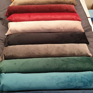 May include: A stack of ten long, rectangular cushions in various colours, including mustard yellow, brown, red, beige, black, grey, green, teal, and blue. The cushions are made of a soft, velvety fabric.