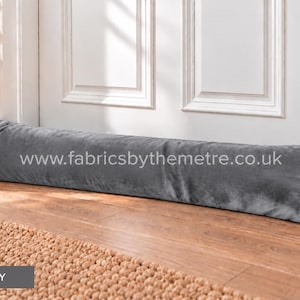 May include: A long, cylindrical grey fabric draught excluder positioned at the base of a white door. The text www.fabricsbythemetre.co.uk is printed in white across the centre of the grey surface. The item has a soft, uniform texture.