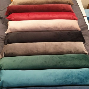 May include: A stack of ten long, rectangular cushions in various colours, including mustard yellow, brown, red, beige, black, grey, green, teal, and blue. The cushions are made of a soft, velvety fabric.