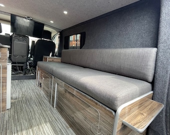 May include: A gray fabric bench seat with a wooden frame and a small wooden shelf. The bench is installed in a van with a wood-look floor and gray walls.