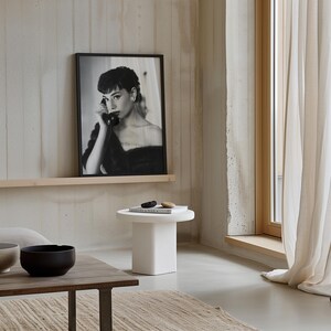 Peut inclure: Une photo en noir et blanc d'une femme au t&eacute;l&eacute;phone est encadr&eacute;e et accroch&eacute;e au mur au-dessus d'une table d'appoint blanche. La table a un livre et deux petites pierres sur le dessus. La pi&egrave;ce a un mur en b&eacute;ton et une fen&ecirc;tre avec des rideaux blancs.