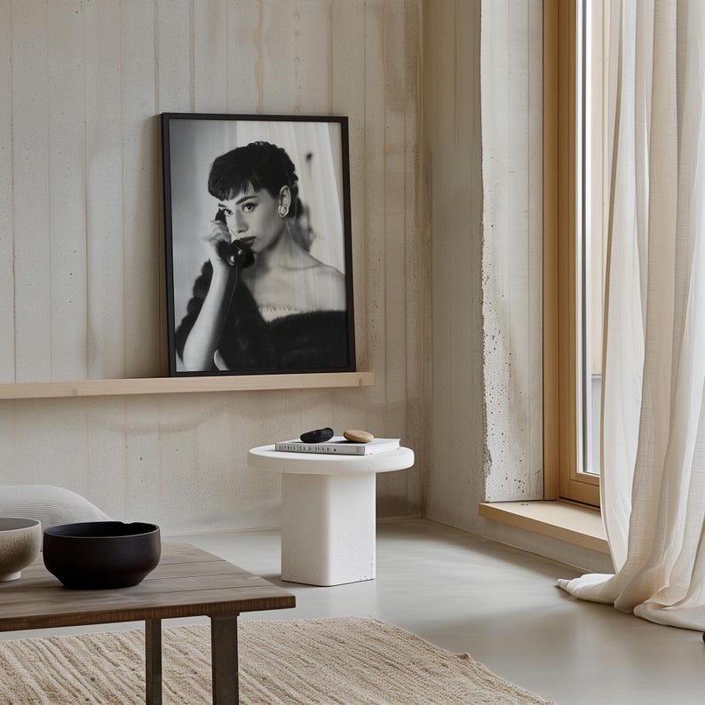 Peut inclure: Une photo en noir et blanc d'une femme au t&eacute;l&eacute;phone est encadr&eacute;e et accroch&eacute;e au mur au-dessus d'une table d'appoint blanche. La table a un livre et deux petites pierres sur le dessus. La pi&egrave;ce a un mur en b&eacute;ton et une fen&ecirc;tre avec des rideaux blancs.
