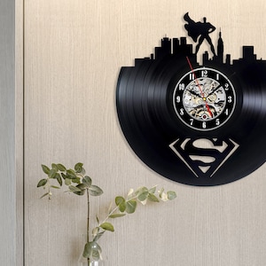 Pode incluir: Relógio de vinil preto com um logotipo do Superman e uma silhueta da cidade. O mostrador do relógio tem algarismos romanos e um mecanismo de relógio visível.