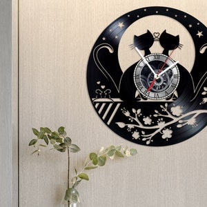 Puede incluir: Reloj de vinilo negro con una silueta de dos gatos con un corazón sobre sus cabezas. La esfera del reloj es un círculo blanco con números romanos negros y un diseño de engranaje negro y plateado. El fondo es negro con un diseño floral blanco.