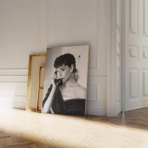 Peut inclure: Photographie en noir et blanc d'une femme en robe noire, tenant un t&eacute;l&eacute;phone rotatif vintage &agrave; son oreille. La photo est encadr&eacute;e d'un cadre dor&eacute; et est appuy&eacute;e contre un mur blanc.