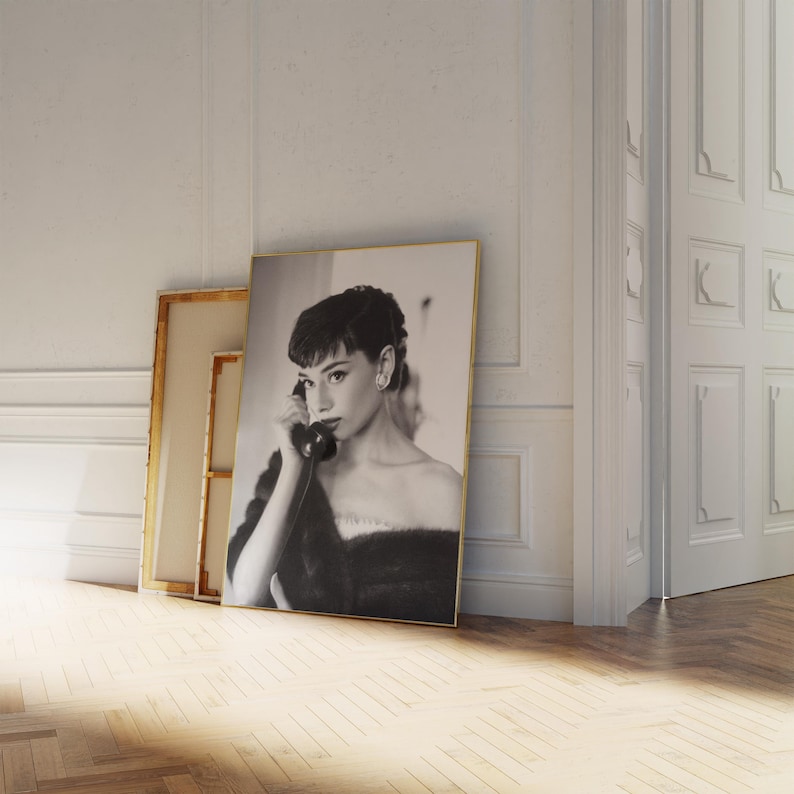Peut inclure: Photographie en noir et blanc d'une femme en robe noire, tenant un t&eacute;l&eacute;phone rotatif vintage &agrave; son oreille. La photo est encadr&eacute;e d'un cadre dor&eacute; et est appuy&eacute;e contre un mur blanc.