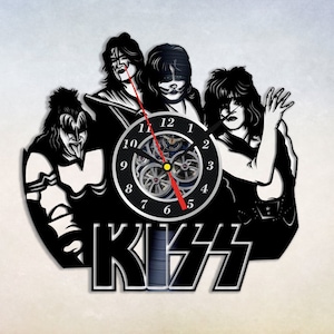 Puede incluir: Reloj de vinilo en blanco y negro con la banda KISS. La esfera del reloj es un círculo negro con números blancos y un mecanismo de reloj plateado. Los miembros de la banda están en silueta en blanco y negro, con el nombre de la banda en letras grandes y negritas debajo de la esfera del reloj.