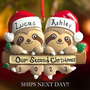 May include: A personalised Christmas bauble featuring two sloths wearing Santa hats with the names "Lucas" and "Ashley" on them. The sloths are holding a wooden sign that reads "Our Second Christmas 2023".
