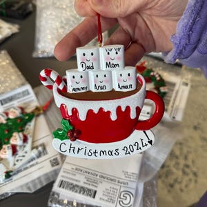 Puede incluir: Un adorno navide&ntilde;o de cer&aacute;mica rojo y blanco con forma de taza de chocolate caliente con malvaviscos. Los malvaviscos est&aacute;n decorados con caras y nombres, incluyendo "Pap&aacute;", "Mam&aacute;", "Sammy", "Arun" y "Aman". La taza tiene un bast&oacute;n de caramelo que sobresale y el texto "Navidad 2021" est&aacute; escrito en la parte inferior.