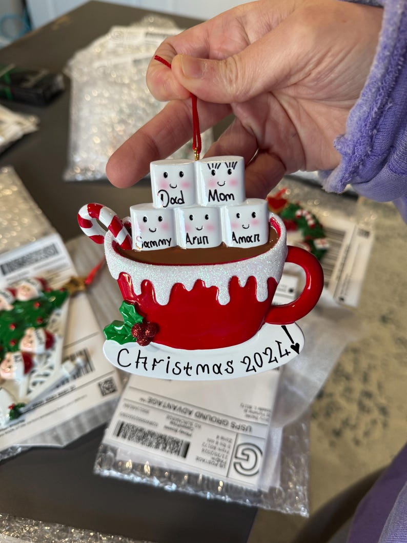 Puede incluir: Un adorno navide&ntilde;o de cer&aacute;mica rojo y blanco con forma de taza de chocolate caliente con malvaviscos. Los malvaviscos est&aacute;n decorados con caras y nombres, incluyendo "Pap&aacute;", "Mam&aacute;", "Sammy", "Arun" y "Aman". La taza tiene un bast&oacute;n de caramelo que sobresale y el texto "Navidad 2021" est&aacute; escrito en la parte inferior.