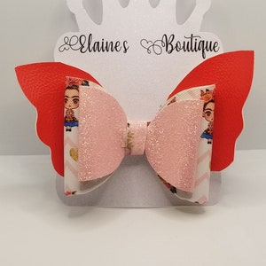 May include: Pink glitter bow with a red faux leather butterfly wing on each side. The bow is decorated with a printed fabric featuring a cartoon image of Frida Kahlo. The bow is displayed on a white crown-shaped stand with the text "Elaines Boutique".