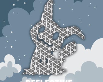 May include: A reflective sticker in the shape of a cartoon ghost, featuring a grey triangular pattern. The word "REFLECTIVE" is written in white at the bottom. The background is blue with clouds and white dots.