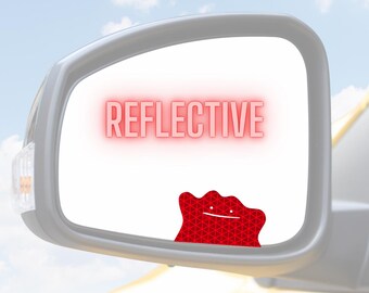 May include: A red reflective decal featuring a blob-like character with two small dots for eyes and a simple line for a mouth, applied to a car side mirror. The word REFLECTIVE is printed in red text above the character on the mirror surface.