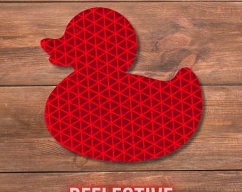 May include: A red rubber duck silhouette featuring a repeating geometric triangular pattern across its surface. The material has a distinct reflective quality, and the word REFLECTIVE is printed in bold, light-coloured capital letters below it.