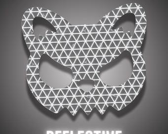 May include: A reflective sticker shaped like a stylised cat head with pointed ears and a triangular nose. The surface features a repeating white geometric triangle pattern on a dark grey background, with the word REFLECTIVE printed below.