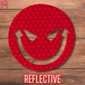 May include: A red reflective sticker with a smiling face with a mischievous grin. The sticker has a triangular pattern and the word "REFLECTIVE" is printed below it.
