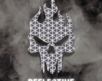 May include: A silver reflective decal shaped like a skull with flames rising from the top. The surface features a repeating geometric triangle pattern throughout. Below the skull, the word REFLECTIVE is printed in bold white capital letters.