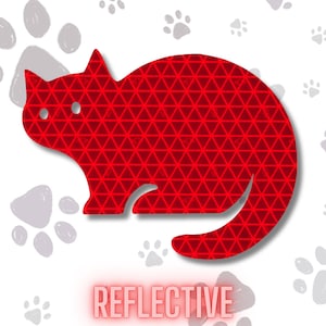 May include: A red cat-shaped sticker featuring a repeating triangular geometric pattern across its surface. The sticker has two circular cutouts for eyes and the word REFLECTIVE printed in red capital letters at the bottom of the design.