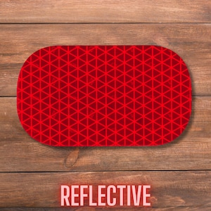 May include: A red oval-shaped reflective safety marker featuring a repeating geometric pattern of small triangles across its entire surface. The object has a smooth, flat finish and is designed with a high-visibility grid structure for light reflection.