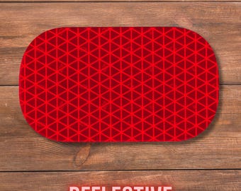 May include: A red oval-shaped reflective safety marker featuring a repeating geometric pattern of small triangles across its entire surface. The object has a smooth, flat finish and is designed with a high-visibility grid structure for light reflection.