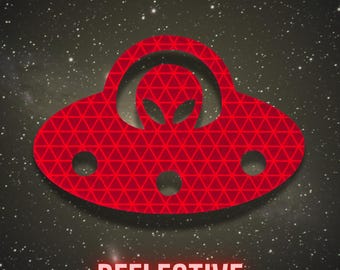 May include: A red UFO graphic with a geometric triangle pattern and a central alien silhouette. The design features three circular cutouts along the bottom edge. Below the graphic, the word REFLECTIVE is printed in a bold, sans-serif font.