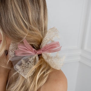 Puede incluir: Un lazo para el pelo rosa y dorado con un delicado diseño de encaje. El lazo está atado con una cinta rosa y tiene un aspecto suave y femenino.
