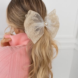 Puede incluir: Un gran lazo de encaje blanco y dorado con un acento de perla. El lazo está atado alrededor de una coleta.