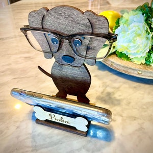 May include: Wooden dog phone stand with glasses holder. The dog is wearing glasses and has a bone-shaped base with the name "Boba" engraved on it.