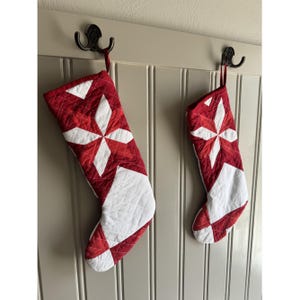 May include: Two quilted Christmas stockings in red and white fabric featuring a geometric star pattern. The stockings are constructed with a diamond-stitched texture and include a fabric loop at the top for hanging on a wall-mounted hook.