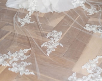 May include: A white bridal veil with delicate lace trim. The veil is made of soft tulle and features a floral pattern. The veil is laid out on a wooden floor.