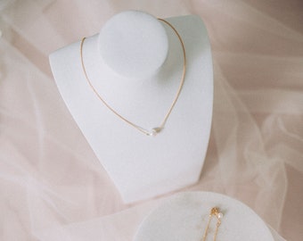 May include: A delicate gold chain necklace with a single white pearl pendant. The necklace is displayed on a white mannequin bust. A matching gold chain bracelet with a single white pearl charm is also shown on a white marble surface.