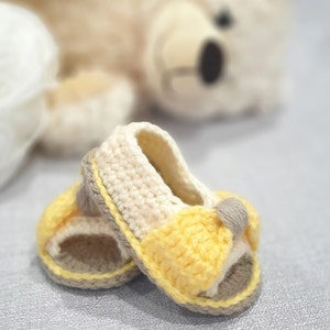 Pode incluir: Um par de sand&aacute;lias de beb&eacute; de croch&ecirc; em amarelo, branco e castanho. As sand&aacute;lias t&ecirc;m uma tira na parte superior e s&atilde;o abertas.