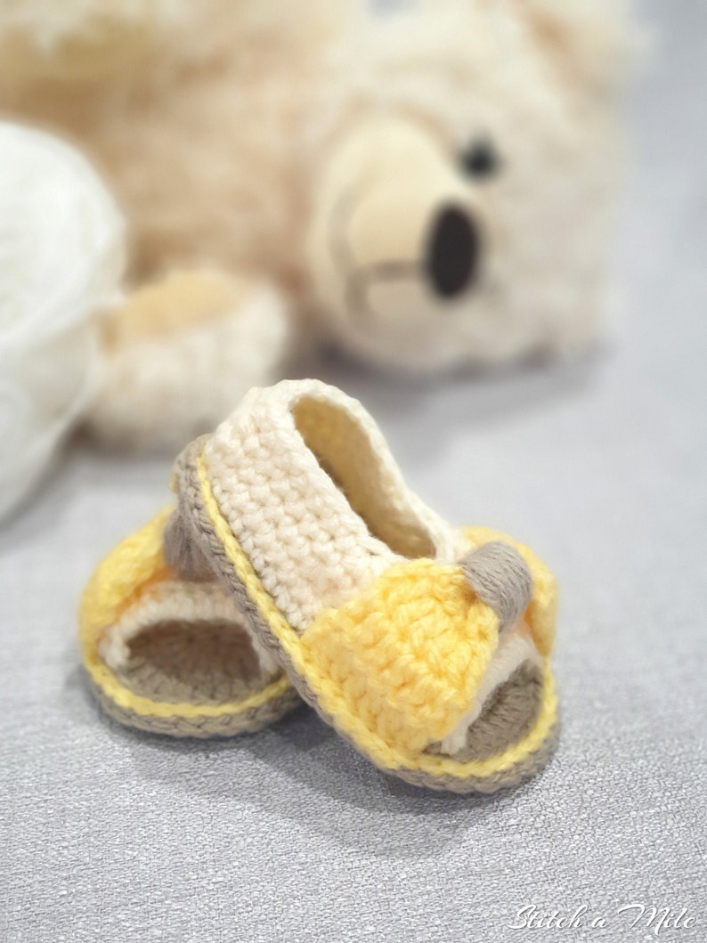 Pode incluir: Um par de sand&aacute;lias de beb&eacute; de croch&ecirc; em amarelo, branco e castanho. As sand&aacute;lias t&ecirc;m uma tira na parte superior e s&atilde;o abertas.