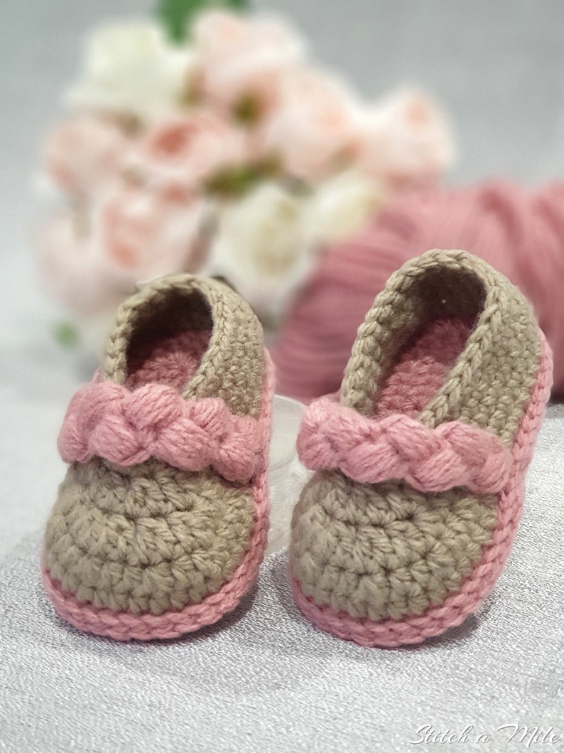 Pu&ograve; includere: Un paio di scarpine per neonati all'uncinetto in beige e rosa. Le scarpine hanno un cinturino rosa intrecciato sulla parte superiore e un bordo rosa attorno al bordo.