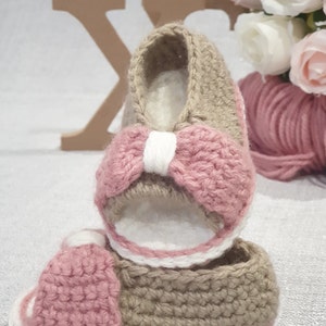 Pode incluir: Um par de sand&aacute;lias de beb&eacute; de croch&ecirc; em rosa e castanho. As sand&aacute;lias t&ecirc;m um la&ccedil;o branco na frente e uma borda branca.
