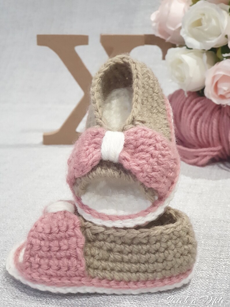 Pode incluir: Um par de sand&aacute;lias de beb&eacute; de croch&ecirc; em rosa e castanho. As sand&aacute;lias t&ecirc;m um la&ccedil;o branco na frente e uma borda branca.